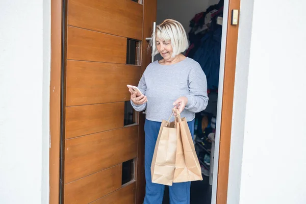 Home delivery. 50s Woman with smartphone opening house entrance door while meeting courier with online order, postwoman delivering package to customer. — Photo