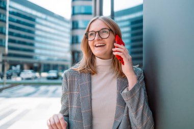 Çekici genç iş kadını şık kıyafetler giyiyor ve açık havada akıllı telefon kullanıyor, ofis binasının yakınında çalışıyor.