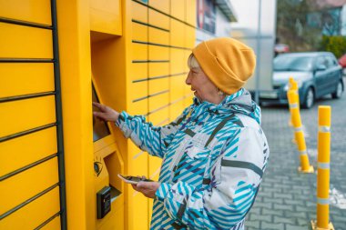 50s 60s Caucasian woman send or receive parcel with self service post terminal machine. Female hand with package