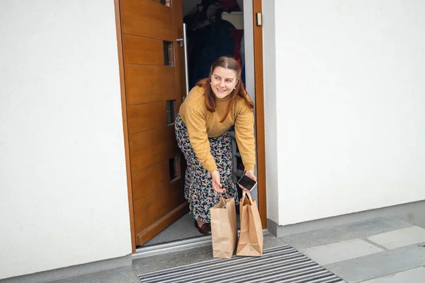 Food delivery. Caucasian woman taking delivered shopping brown paper bag from floor near a home, checking online purchases with a smart phone — Photo