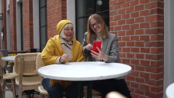 Beautiful young girl and 50s woman using digital smartphone in coffee shop while spending time together on the cafe terrace — Stok Video