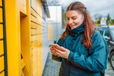 Beyaz kadın akıllı telefon ve akıllı dağıtım uygulaması gösteriyor. Sarı otomatik posta terminalinin yanında.. 