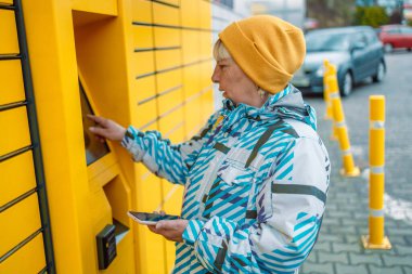 50s 60s Caucasian woman send or receive parcel with self service post terminal machine. Female hand with package