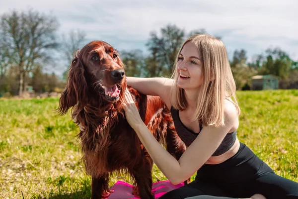 Cute young caucasian woman is meditating while sitting on a rug on the lawn on a sunny summer with Irish Setter dog. — Foto Stock