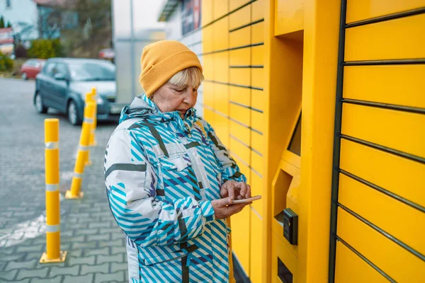 Caucasian woman showing smartphone with smart delivery app near a yellow automatic post terminal. — Foto de Stock