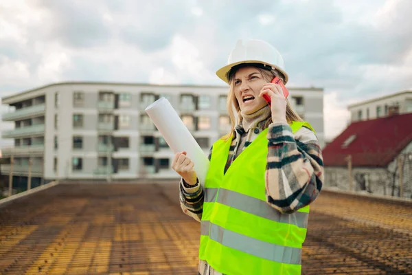 Angry female construction worker in hardhat with smartphone outdoors checking construction. Industry concept