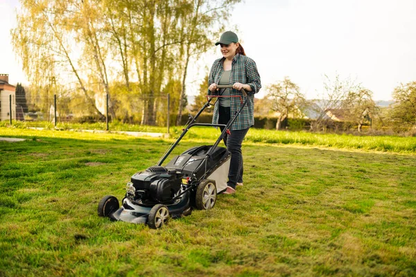 Pushing grass trimming lawnmower. caucasian 30s woman using electric lawn mower while working at garden. — Stockfoto