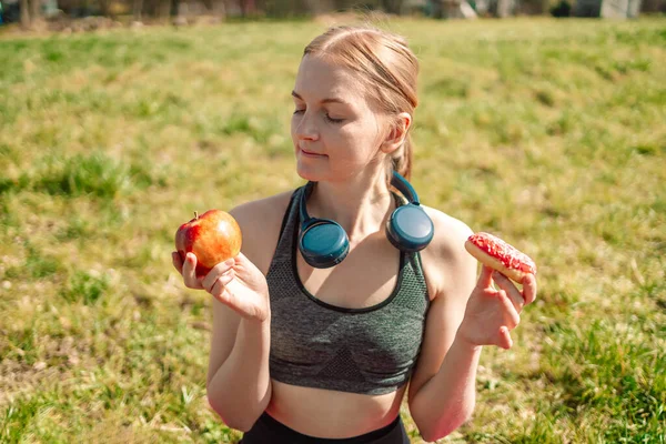 Wahl zwischen Apfel und Donut. Ernährungskonzept. Das blonde Mädchen hält einen rosafarbenen Donut und einen Apfel in der Hand. Süßigkeiten sind ungesundes Junk Food. Diäten. Gesunde Ernährung. — Stockfoto