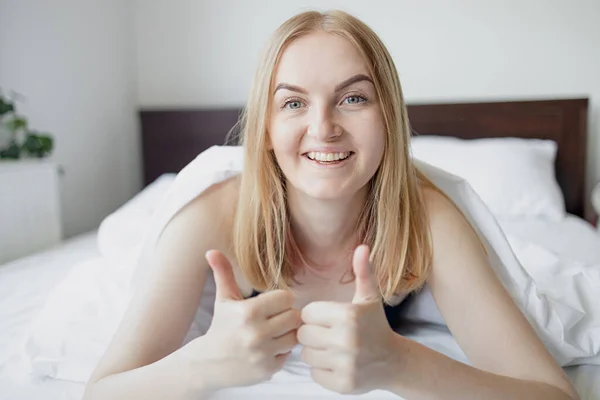 Feliz joven rubia de 30 años mujer muestra como con la mano tendida en la cama cubierta con ropa de cama blanca temprano en la mañana. —  Fotos de Stock