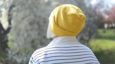 Şık giyinmiş, sarı şapkalı, mutlu ve sarışın bir kadının portresi. Baharda parkta tek başına yürürken kameraya gülümsüyor.