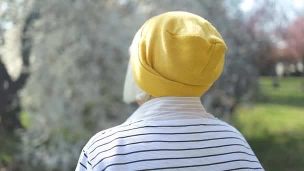 Πορτρέτο μιας όμορφης ξανθιάς ηλικιωμένης γυναίκας με κομψά ρούχα και κίτρινο καπέλο που χαμογελά στην κάμερα περπατώντας μόνη της στο πάρκο την άνοιξη — Αρχείο Βίντεο
