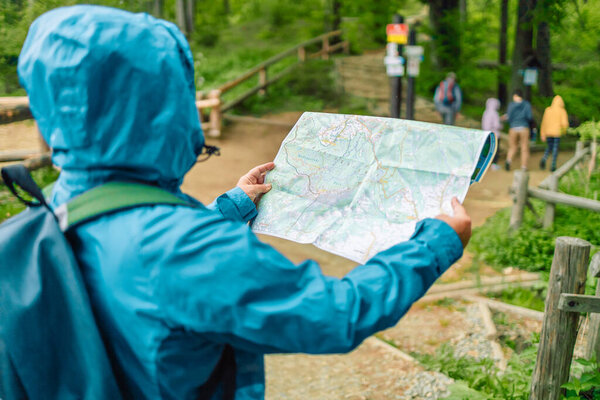 Hiking young woman traveler with backpack checks map to find directions on top of mountain with nature background