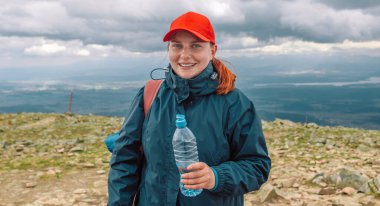 Çantalı genç Avrupalı kadın dağın tepesinde bir plastik su gösteriyor.