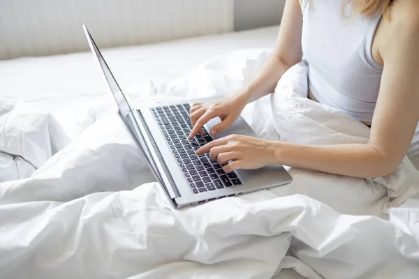 Happy casual beautiful woman working on a laptop sitting on the bed at the morning in the house. Working at home, freelance concept.