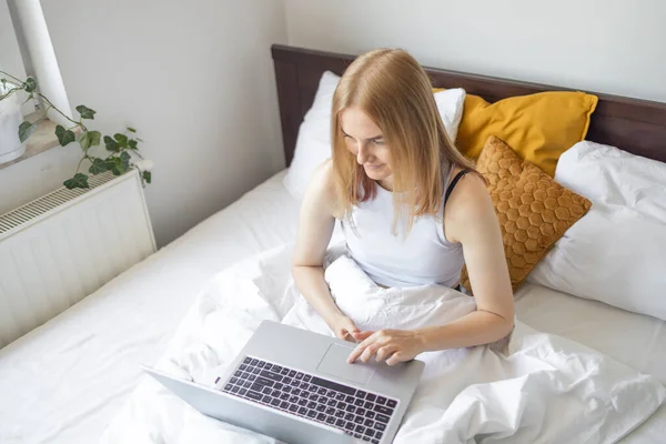 Happy casual beautiful woman working on a laptop sitting on the bed at the morning in the house. Working at home, freelance concept.