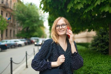 Açık havada gülümseyen sarışın bir kadının portresi. Parktaki akıllı telefondan mutlu bir şekilde konuşuyor..