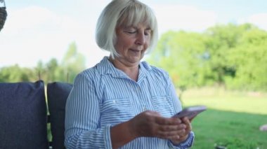 Attractive blonde 50s 60s woman of retirement age sitting on park bench making video call, looking at smartphone camera. Granny retired uses modern gadget internet talking on mobile phone outdoor