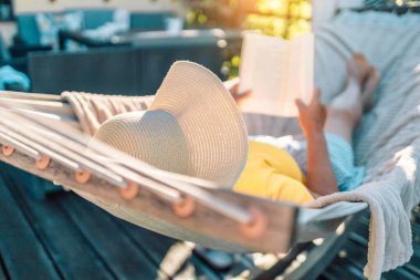 Beautiful woman with hat reading book in comfortable hammock at green garden. 