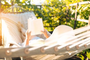Beautiful woman with hat reading book in comfortable hammock at green garden. 