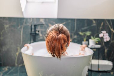 Back view shot of young woman lying in bathtub with her eyes closed. Female relaxing in bathtub. 