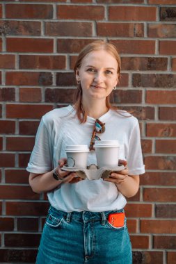 Neşeli beyaz kadın portresi gülümsüyor ve şehir caddesinde yürürken iki tane kağıt kahve fincanı tutuyor.. 