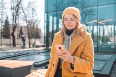Şehir parkında akıllı telefon kullanan mutlu bir kız. Şehir merkezindeki bankta oturuyor.