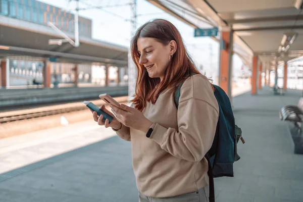 Pasaportlu genç kızıl saçlı kadın istasyonda bekliyor ve akıllı telefon kullanıyor. Demiryolu taşımacılığı konsepti. Hızlı trenle tatile git.. 