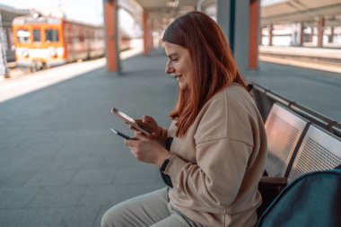 Pasaportlu genç kızıl saçlı kadın istasyonda bekliyor ve akıllı telefon kullanıyor. Demiryolu taşımacılığı konsepti. Hızlı trenle tatile git.. 