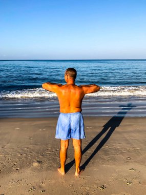 Bir adam spor yapmaya gider, deniz kıyısında egzersiz yapar.