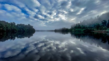 Vivid cloud reflections on the surface of the tranquil lake in the early morning mist and fog