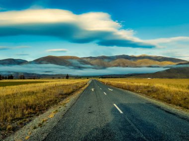 Asfalt yol, tepelerin üstünde ve altında kabarık bulutlara doğru gidiyor. 