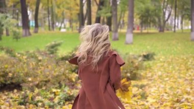 Genç ve güzel Wonam sonbahar parkında yürüyüş yapıyor, koşuyor ve yaprakları fırlatıyor. Yol boyunca koşan genç ve mutlu bir modelin açık hava çekimi. Renkli görüntü. Yüksek kalite FullHD video
