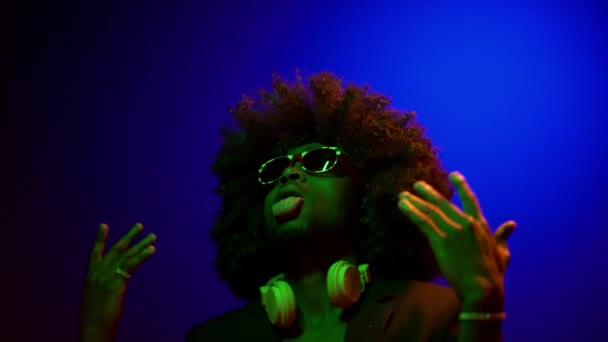 Afro American DJ en Neons muestra la lengua y el baile sobre un fondo aislado. En una peluca, una pluma joven y gafas mueven rítmicamente sus manos — Vídeo de stock