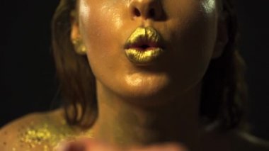 Golden Woman. Female Gold Lips Blow Gold Glitter from Hands. Beauty Fashion Lady with Golden Make-up, Gold Skin, Close-up. Dark Background