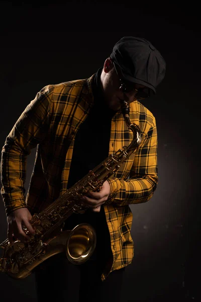 Stylish Tenor Sax Player in Yellow Plaid Shirt, Cap and Sunglasses Plays Saxophone on Black Background. Saxophonist Plays Jazz Music with Sax Alto. Close-up