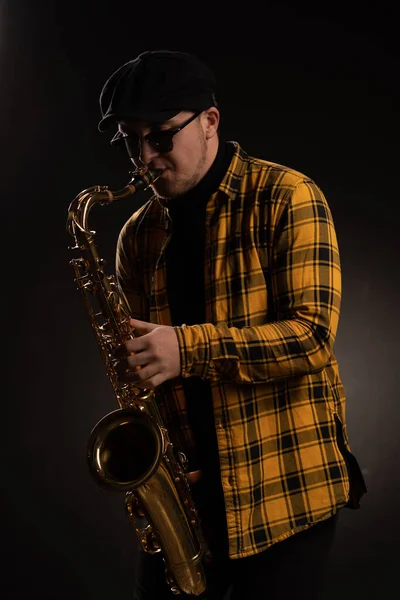 Homem Saxofonista em Camisa Amarelo Checkered, Boné e Óculos de Sol Elegantes Segurando um Saxofone Alto. Fundo preto. Retrato de close-up — Fotografia de Stock