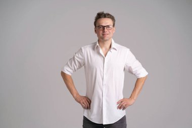 Beyaz gömlekli, gözlüklü, kalçalı, gülümseyen ve beyaz arka planda kendine güvenen yakışıklı bir adamın portresi.