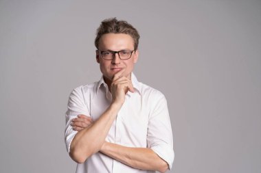 Beyaz gömlekli ve gözlüklü genç, yakışıklı ve arkadaş canlısı bir adam gülümsüyor, mutlu ve kendinden emin bir ifadeyle kollarını beyaz bir arka plan stüdyo çekiminde kavuşturuyor.