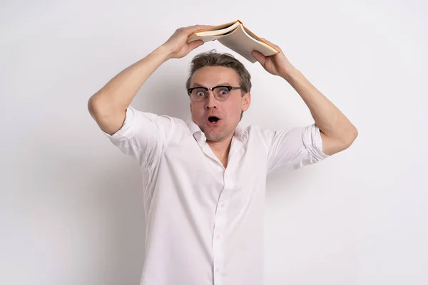 Funny man in glasses hiding under book holding it above head with amazement emotion on his face wear white shirt and grey jeans isolated white background. learning concept — Stock Photo, Image