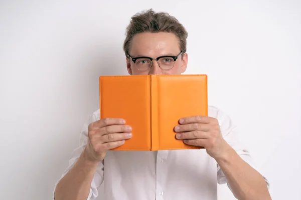 Man hide behind orange notebook or journal in eye glasses wearing white shirt isolated on white background or in the office. Portrait of a shy young businessman holding his diary — Stock Photo, Image