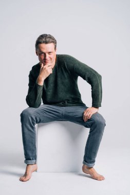 Beyaz küpte oturan yakışıklı çıplak ayakçı, eli çenesine değmiş, kameraya bakıyor, siyah kot pantolon giyiyor ve beyaz arka planda yeşil-gri kazak örüyor.