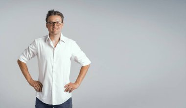 Beyaz gömlekli ve gözlüklü kalçalı yakışıklı serbest bir adam, beyaz bir arka planda izole edilmiş yüzünde kendine güvenen ve gülümseyen bir pozitif ifade. Boşluğu kopyala