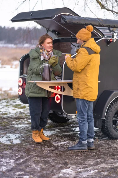 Standing near camper kitchen mature couple enjoying hot coffee during journey winter camper vacation, man and woman with cups in hands. Family spend time camping, journey together.