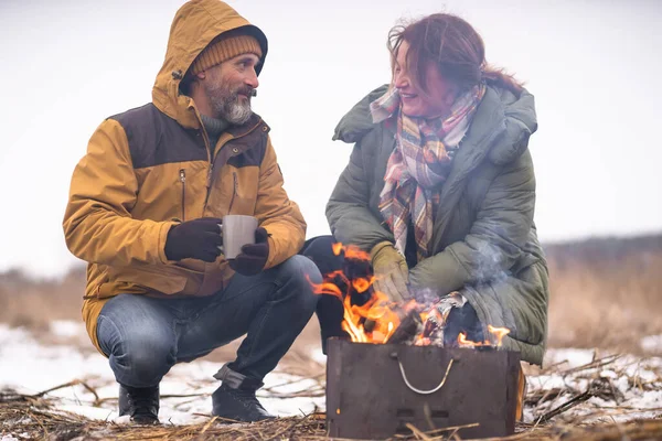 在篝火边热身 中年男人和女人在野外喝着热茶或咖啡 一对成熟的夫妇在冬季森林篝火边烧烤 家庭旅行 家庭假概念 — 图库照片