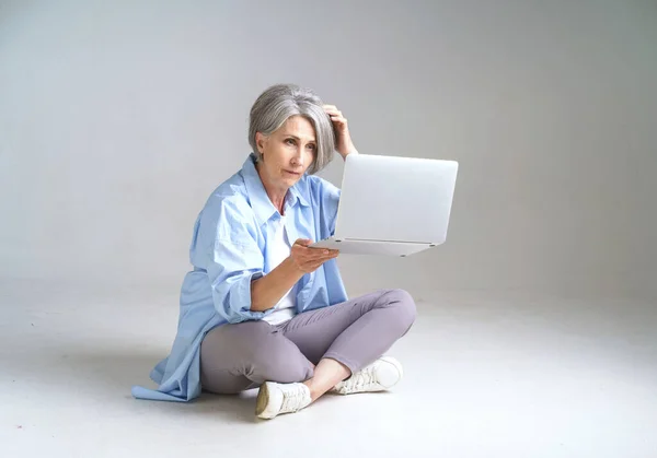 Frustrated mature woman holding laptop in hand scratching her head looking at it sitting on the floor isolated on white background. Mature people and technologies. Senior woman with laptop.