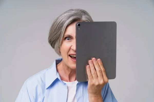 Angry mature grey haired woman hide behind digital tablet posting bad comments or trolling other users online in social media. Senior woman in blue shirt isolated on white background.