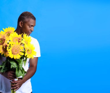 Romantic young African American guy, man holding bouquet of sunflowers with head turned sideways down wearing white t-shirt isolated on blue background. Romantic young man with flowers.