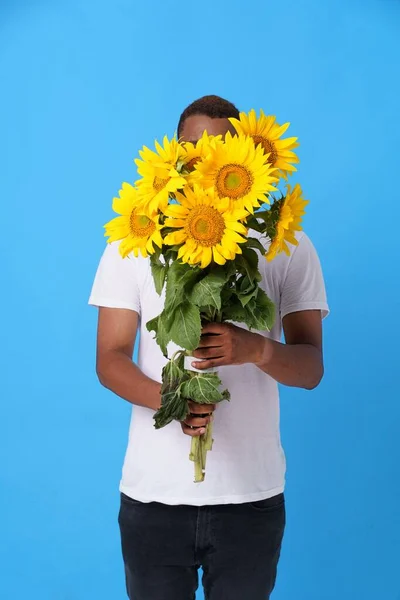 Hide behind bouquet of sunflowers young African American man wearing white t-shirt and isolated on blue background. Romantic young man with flowers. Man with summer flowers. No face visible.