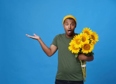 Amazed, shocked young African American man with armful of sunflowers pointing with hand up wearing green t-shirt and yellow hat isolated on blue background. Flower delivery advertisement concept. 