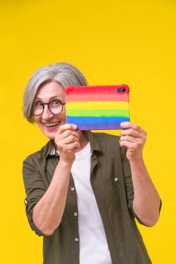 Looking out behind digital tablet in colorful rainbow silicone cover, case mature woman isolated on yellow background. Digital tablet in woman hands supporter human rights, LGBTQ support concept.